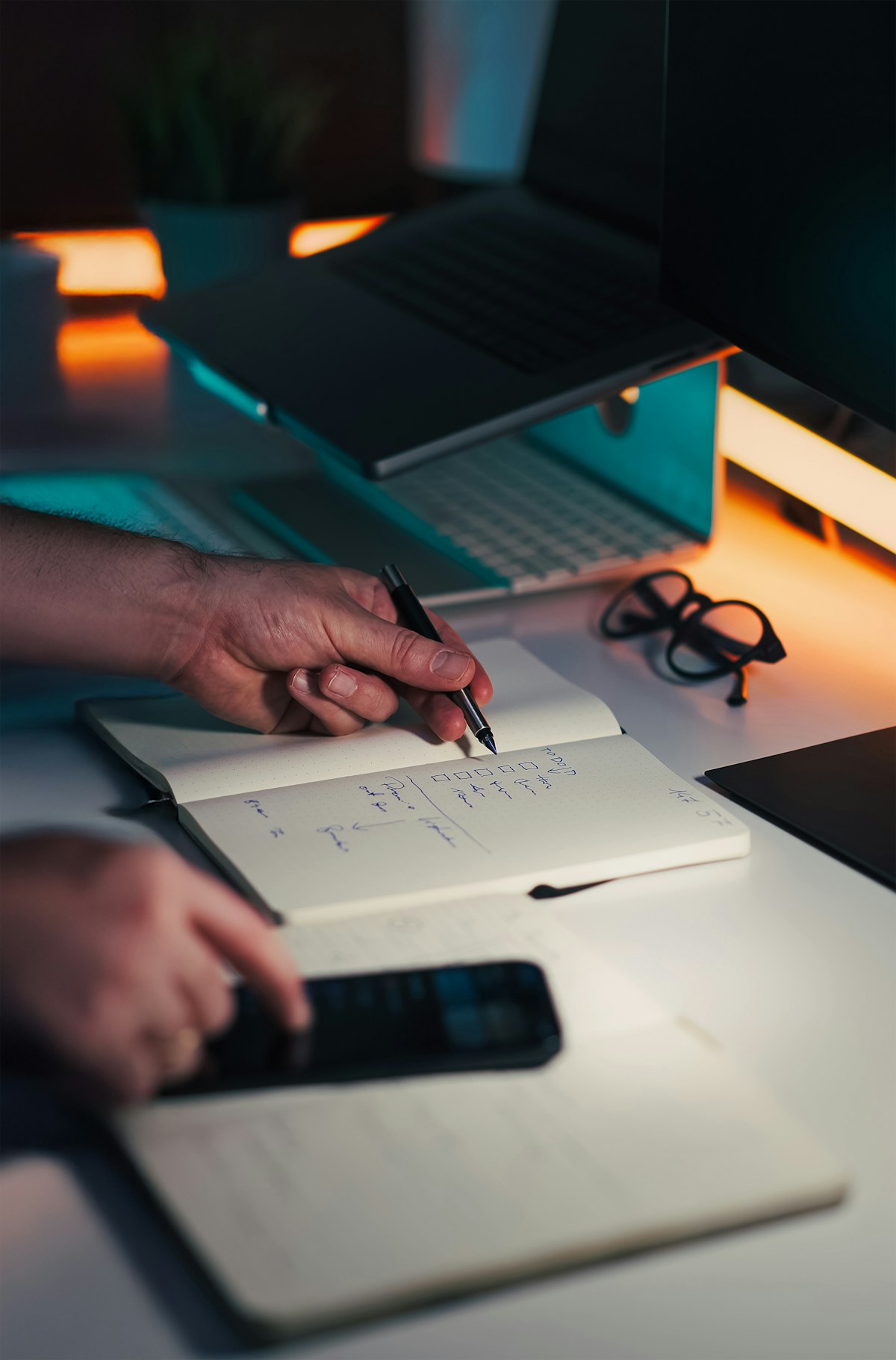 Hands taking notes in a notebook beside a laptop and phone.
