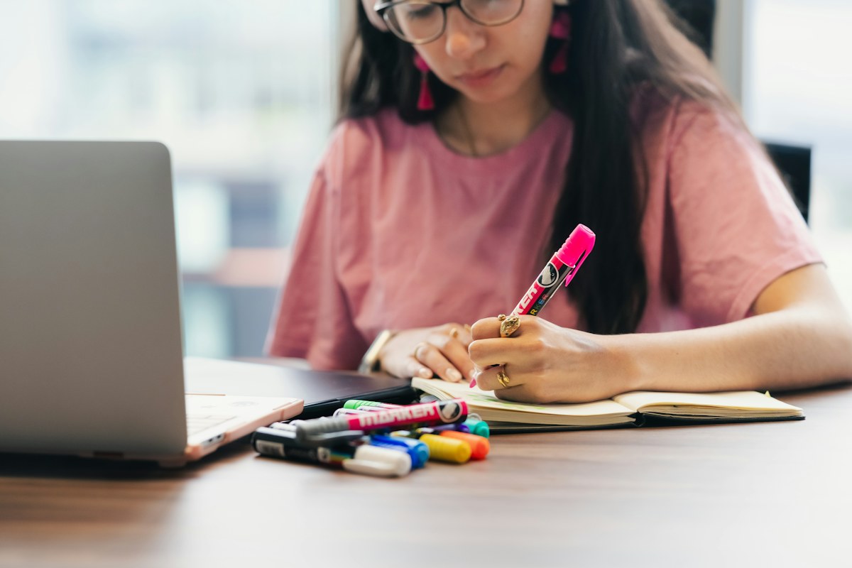 A woman making notes in a notebook beside her laptop.