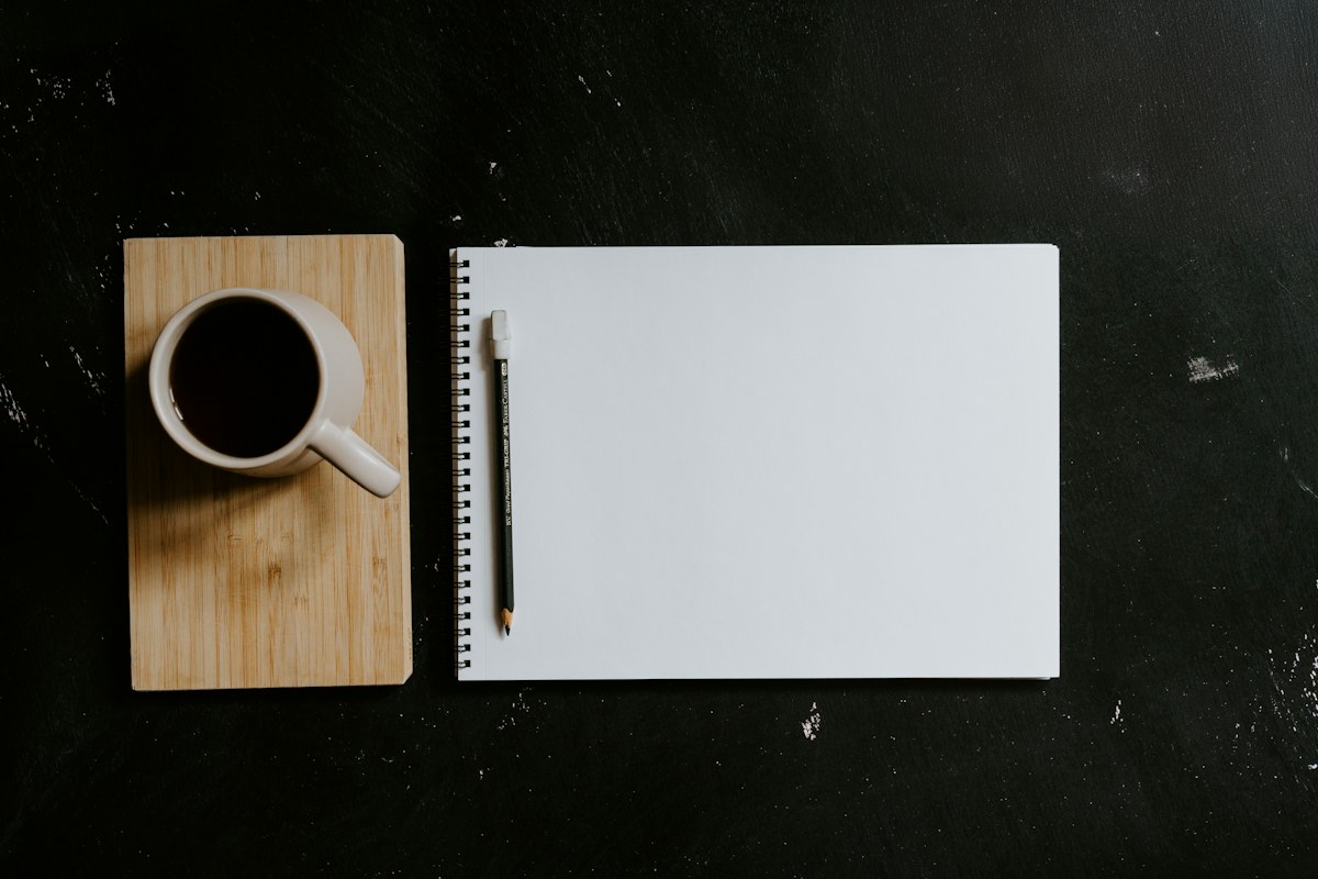 A blank notebook and pencil beside a cup of coffee on a dark desk.